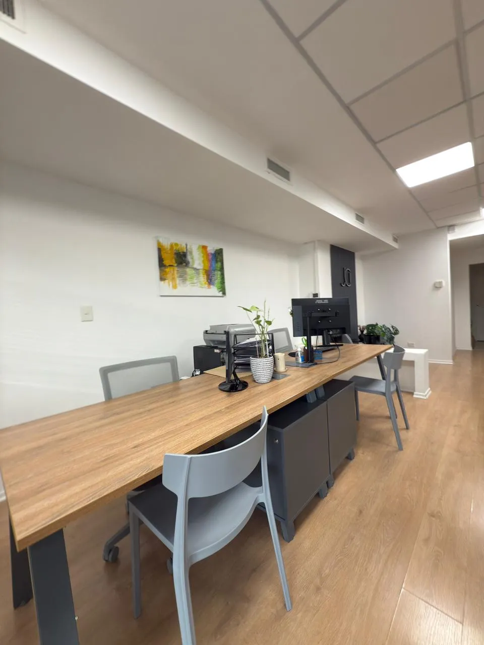 Escritorio compartido de madera con monitores y sillas ergonómicas grises en un ambiente de oficina abierto.